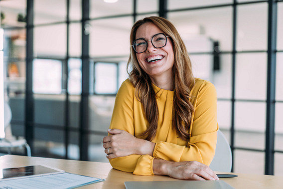 A smiling woman wearing glasses and a yellow blouse sits at a desk in a modern office with glass walls. She rests her arms on the table near documents and a laptop, conveying a relaxed, friendly, and professional mood.