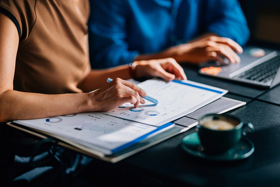 Two colleagues reviewing financial data and charts. The focus is on teamwork and analytical skills in a modern office setting with a laptop and coffee.