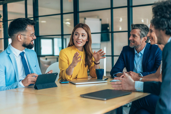 A diverse group of five professionals are seated around a conference table in a modern office. A woman in a bright yellow blouse is speaking animatedly while others listen and engage, suggesting a collaborative discussion or team meeting. Laptops, a tablet, and a coffee mug are on the table.