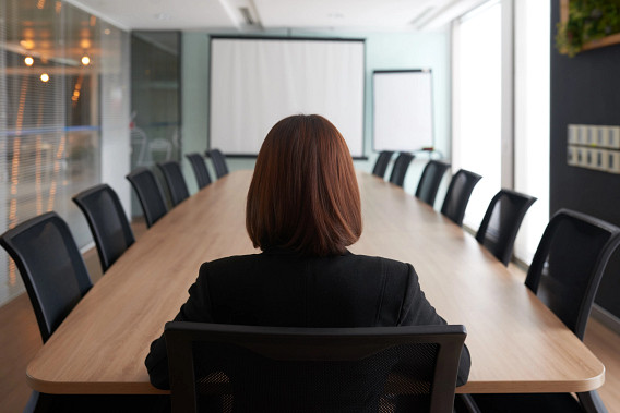 A person sits alone at the head of a long conference table, viewed from behind, in a modern meeting room. Empty chairs line both sides of the table, with presentation screens and large windows creating a quiet, formal atmosphere.