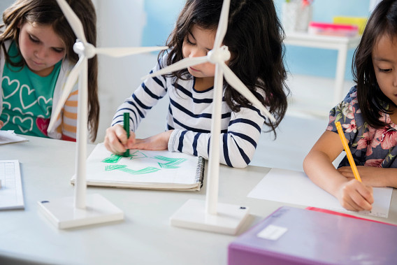 Three elementary school-aged girls engaged in a hands-on educational activity involving a small wind turbine model, suggesting they are learning about renewable energy or environmental topics in a structured classroom setting.