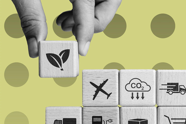 Close-up of a hand placing a wooden cube with a leaf icon on top of stacked wooden blocks showing symbols for transport, CO₂ emissions, fuel, shipping, and shopping, set against a light green background with circular patterns.