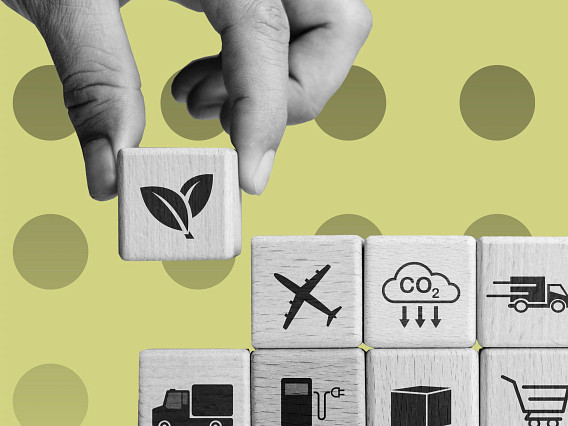 Close-up of a hand placing a wooden cube with a leaf icon on top of stacked wooden blocks showing symbols for transport, CO₂ emissions, fuel, shipping, and shopping, set against a light green background with circular patterns.