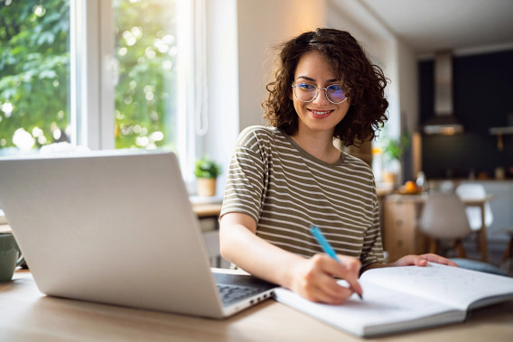 A person with curly hair and glasses sits at a desk, smiling while writing in a notebook. A laptop is open in front of them, and a window with greenery outside is in the background, providing natural light.